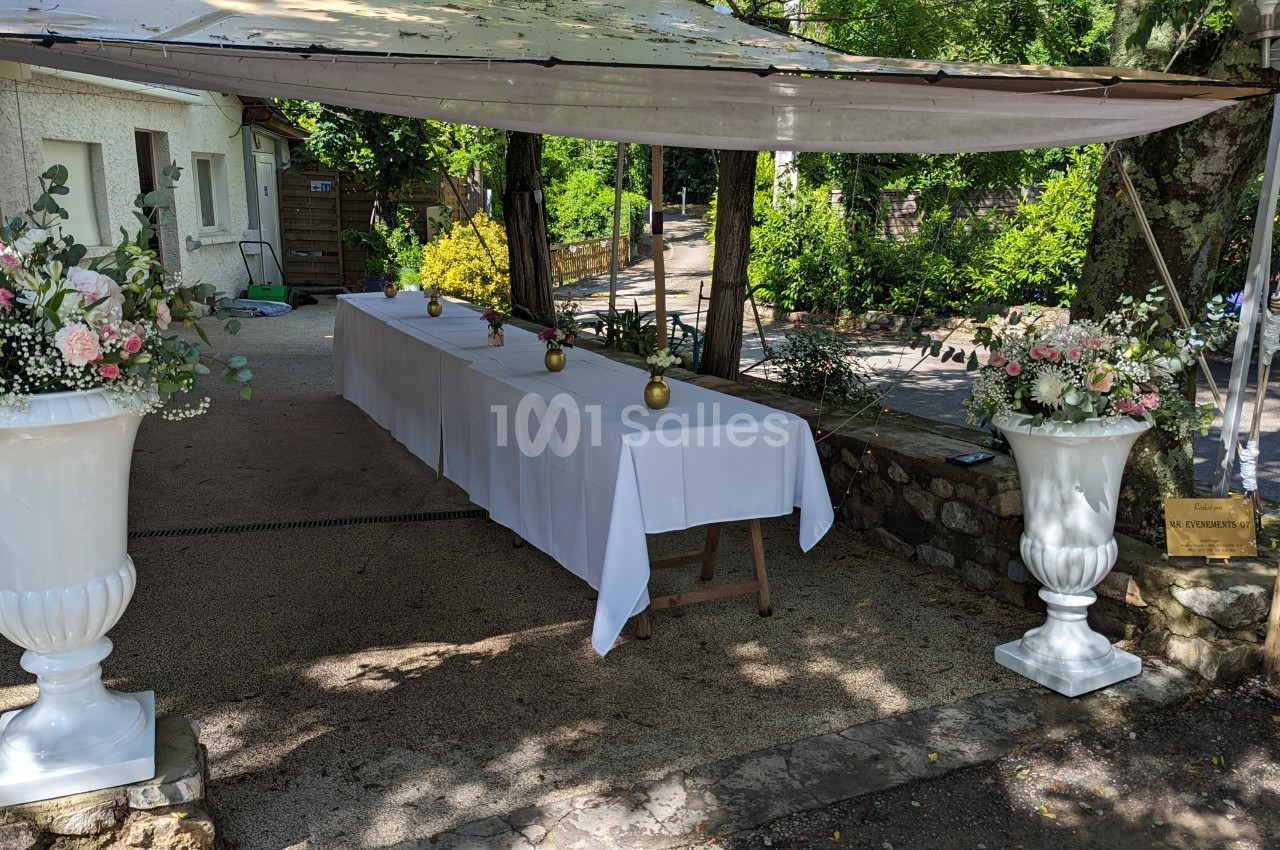 Table décorée avec une nappe blanche sous un auvent, entourée de grands vases fleuris dans un cadre extérieur verdoyant.