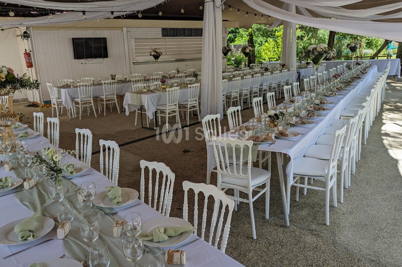 Tables décorées avec nappes blanches, chaises assorties et arrangements floraux dans un espace extérieur couvert.