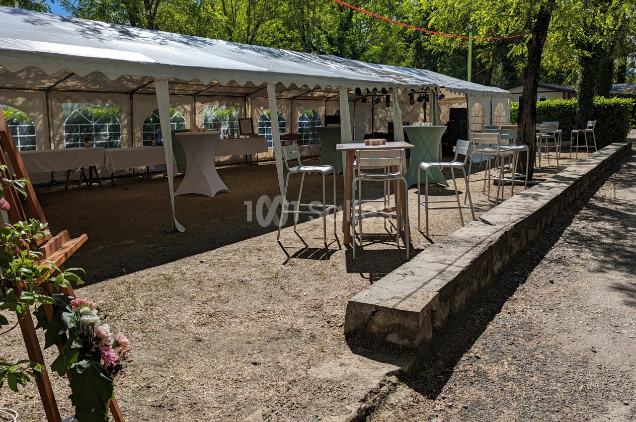 Espace extérieur avec une grande tente blanche, des tables hautes et des chaises, entouré d'arbres sous un ciel ensoleillé.