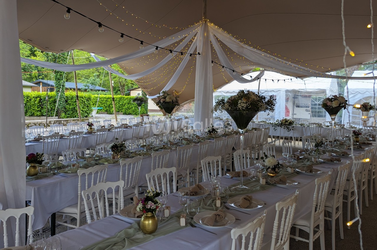 Salle de réception décorée pour un événement, avec longues tables dressées, chaises blanches et guirlandes lumineuses.