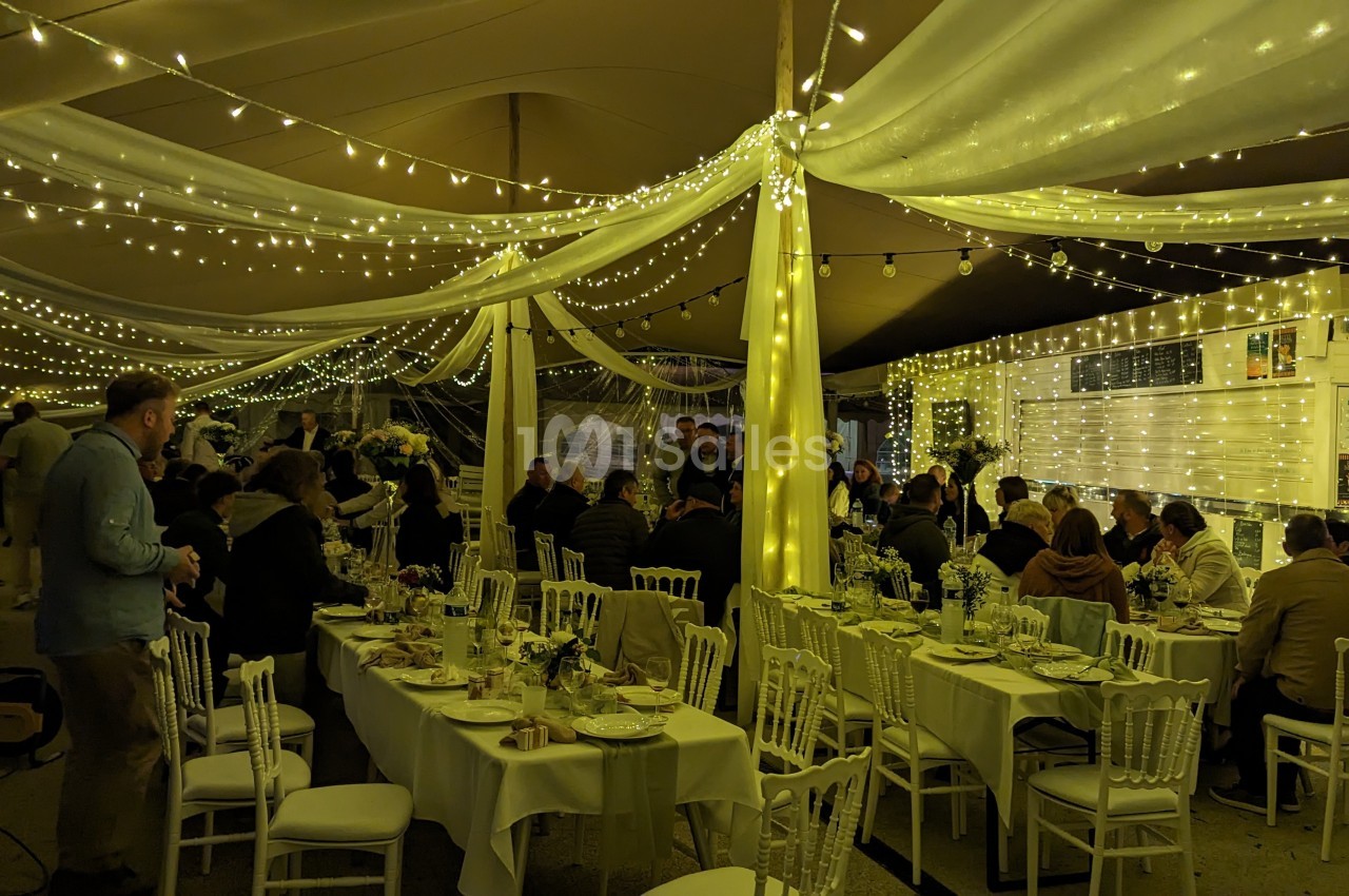 Salle de réception décorée avec des guirlandes lumineuses, tables dressées et invités assis ou debout.