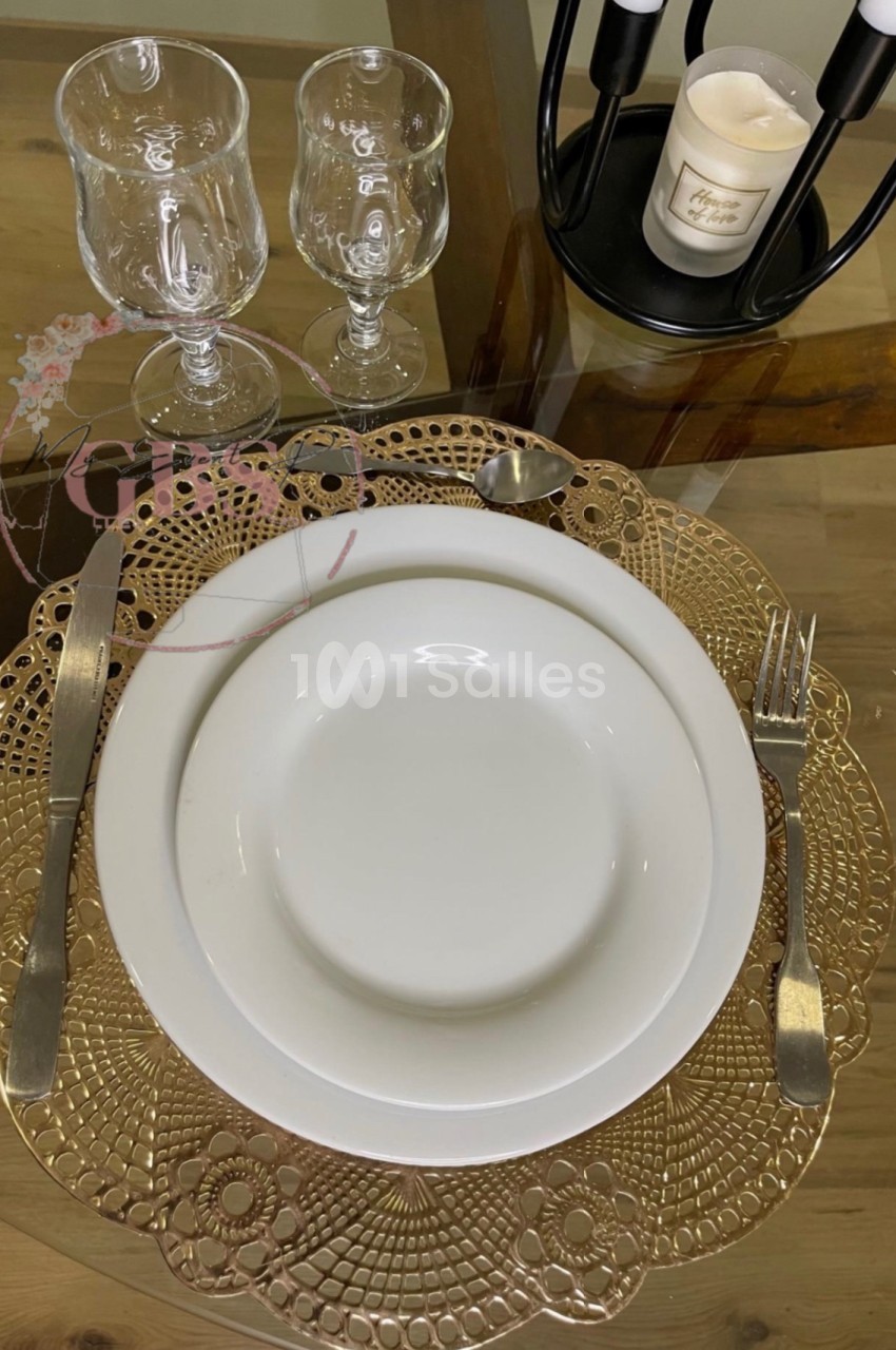Table dressée avec une assiette blanche sur un napperon doré, couverts en métal et verres en cristal.