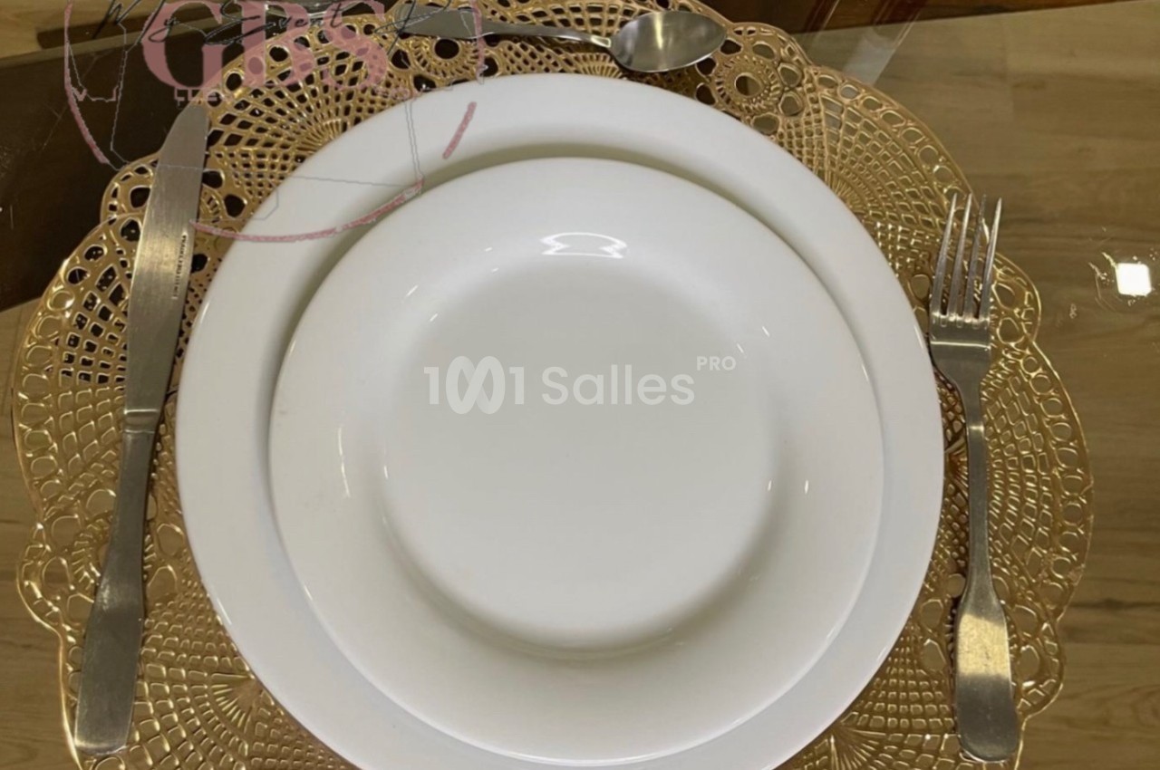 Assiette blanche posée sur un napperon doré, accompagnée de couverts en métal sur une table en verre.