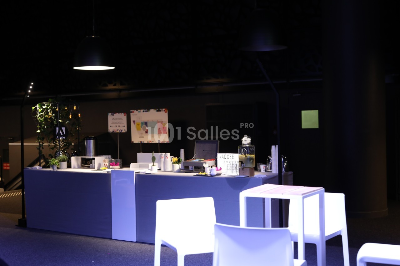 Comptoir moderne avec ordinateur, plantes et accessoires, entouré de chaises blanches dans un espace faiblement éclairé.