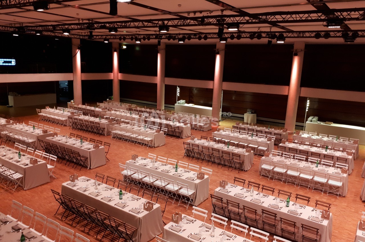 Salle de réception avec de longues tables dressées pour un événement, éclairée par des lumières chaudes.