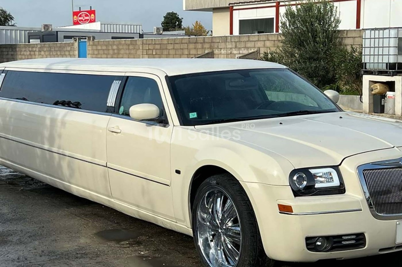 Limousine blanche stationnée sur un sol humide devant un bâtiment et un panneau publicitaire en arrière-plan.