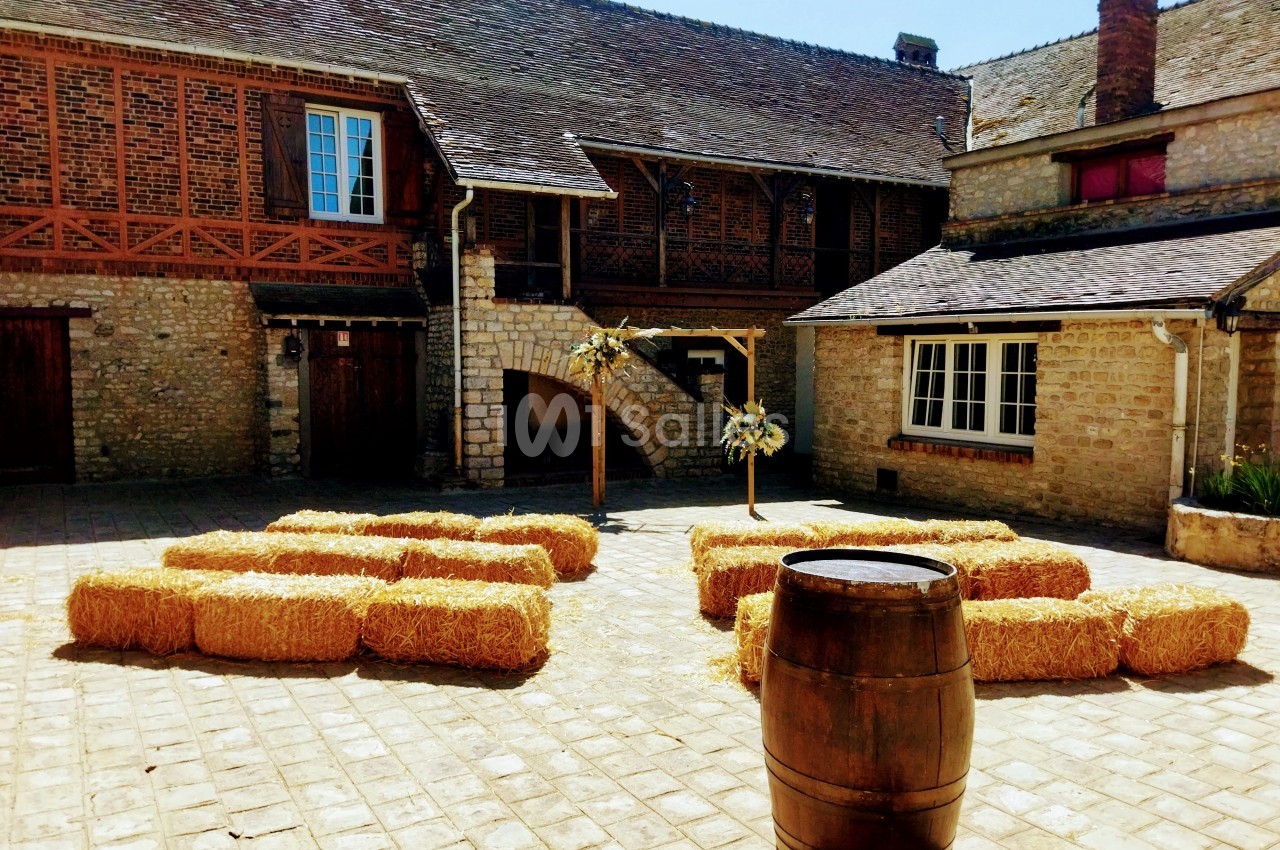 Cour pavée avec des bottes de paille disposées en cercle, entourée de bâtiments en pierre et en bois.