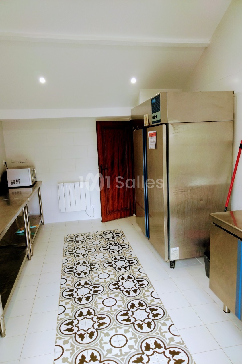 Cuisine professionnelle avec équipements en inox, réfrigérateur, micro-ondes et sol carrelé décoratif.