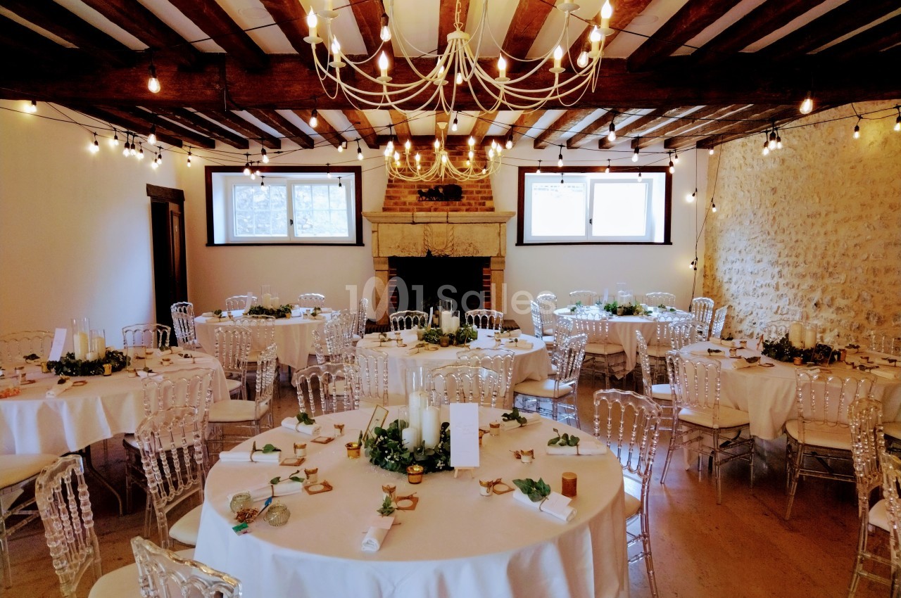 Salle de réception décorée avec des tables rondes, nappes blanches, chaises transparentes et guirlandes lumineuses…