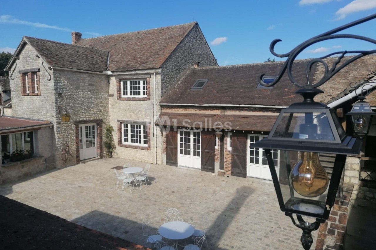 Cour pavée avec tables et chaises blanches, entourée de bâtiments en pierre et éclairée par un lampadaire.