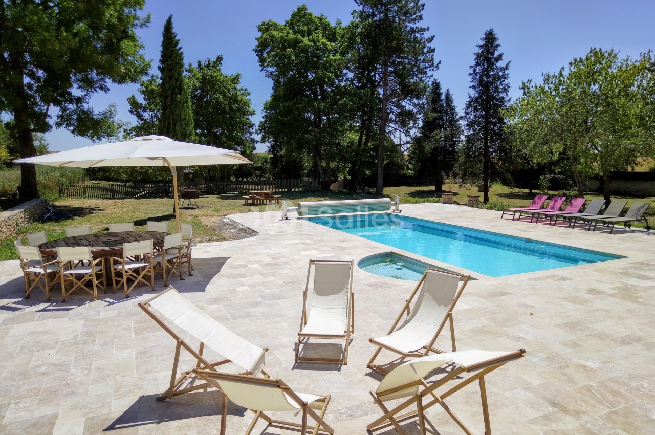 Terrasse avec piscine, transats et parasol, entourée d'arbres dans un jardin ensoleillé.