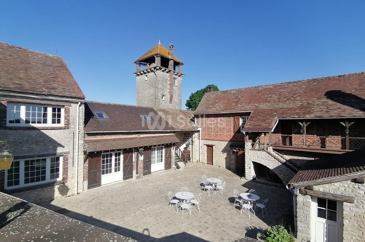 Cour intérieure pavée avec tables et chaises, entourée de bâtiments en pierre et d'une tour avec toit en bois.