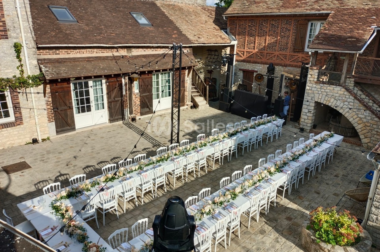 Tables de banquet décorées pour un événement en plein air dans une cour pavée entourée de bâtiments en pierre et en bois.