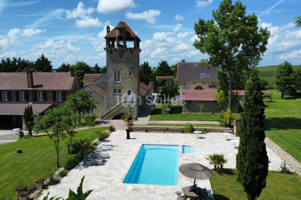 Vue d'un domaine avec une tour en pierre, une piscine entourée de verdure et des bâtiments annexes sous un ciel dégagé.