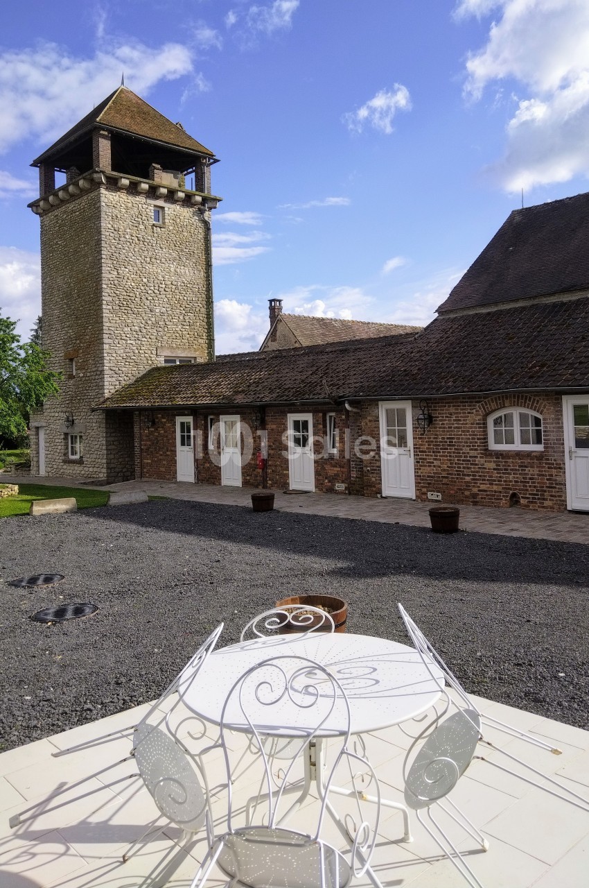 Cour pavée avec une table blanche en fer forgé au premier plan, une tour en pierre et des bâtiments en briques en arrière…