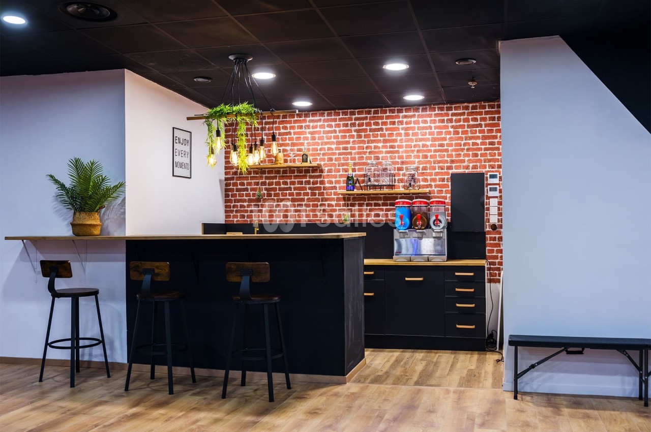 Cuisine moderne avec comptoir noir, tabourets en bois, mur en briques rouges, étagères décorées et plantes suspendues.