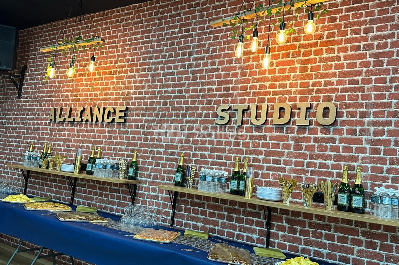 Mur en briques rouges avec étagères contenant des bouteilles, verres et snacks, éclairé par des suspensions lumineuses.