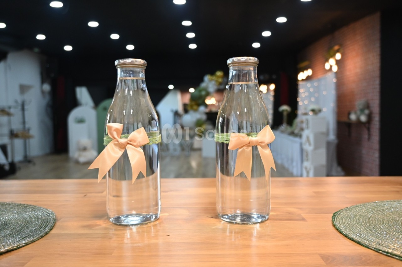 Deux bouteilles en verre décorées de rubans beige et vert posées sur une table en bois dans une salle éclairée.