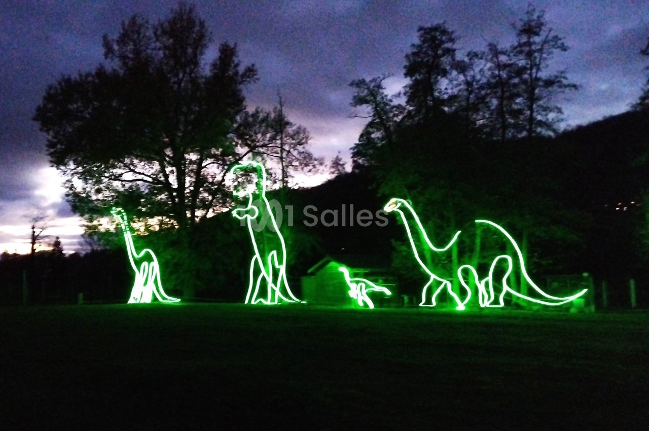 Silhouettes lumineuses de dinosaures en néons verts dans un parc, sur fond de crépuscule et d'arbres.