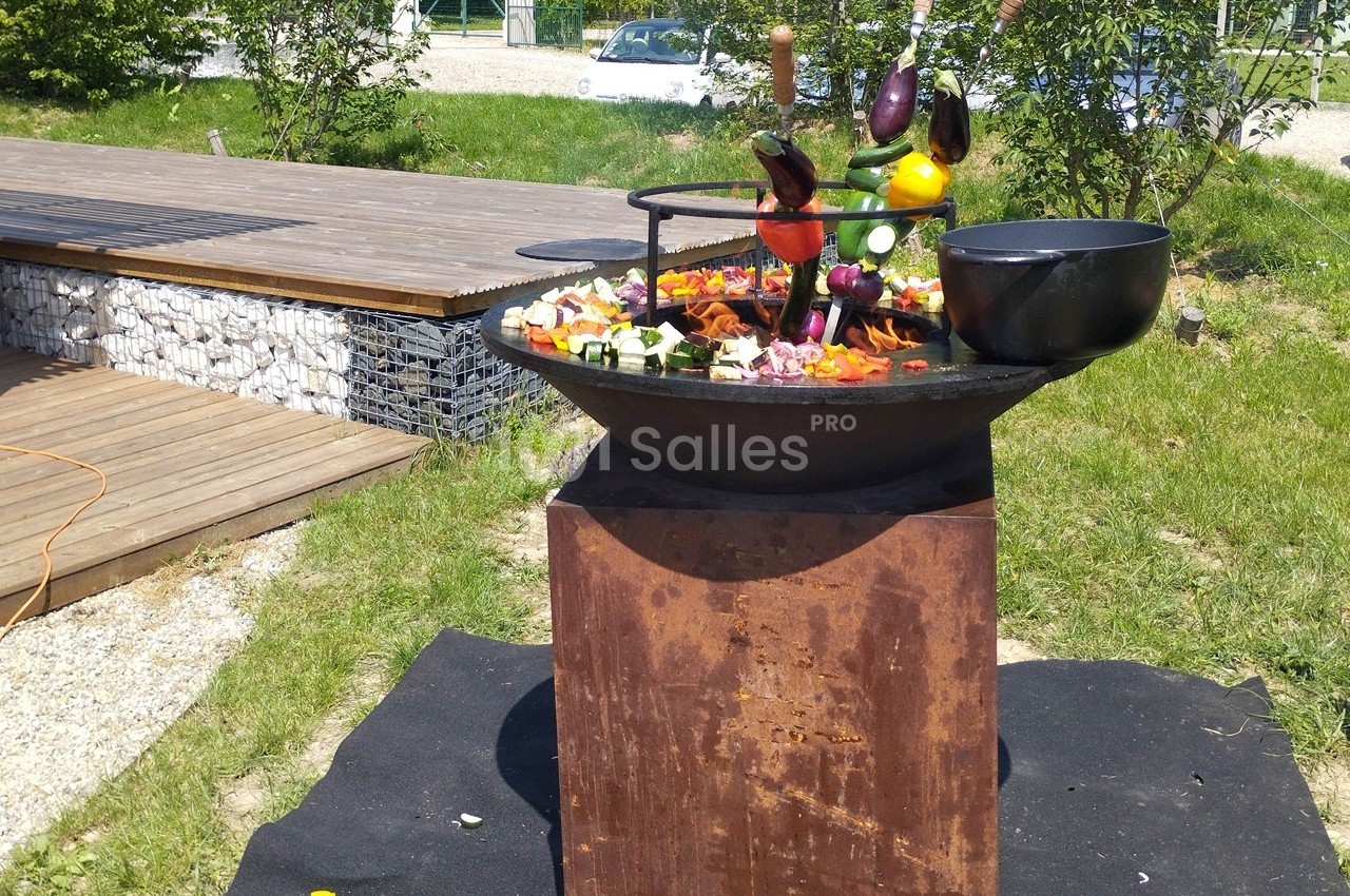 Légumes variés grillant sur une plancha en extérieur, entourée de verdure et d'une terrasse en bois.