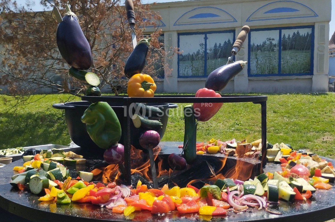 Légumes variés cuits sur un brasero en plein air, avec un bâtiment peint en arrière-plan.