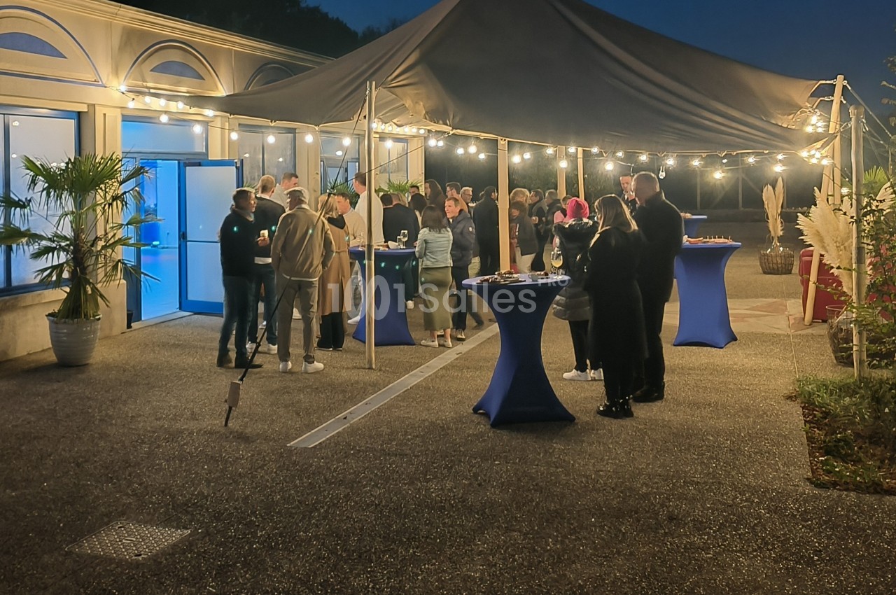 Groupe de personnes réunies sous une tente éclairée en soirée, avec des tables hautes et un décor extérieur.