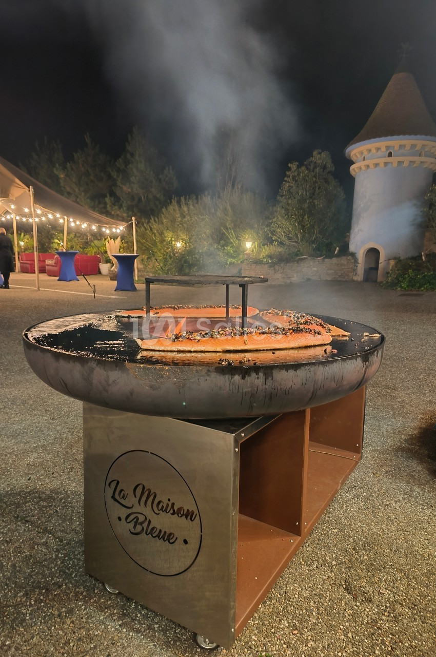 Barbecue circulaire en extérieur, avec des filets de saumon en cuisson, devant un décor nocturne et une tour en arrière-plan.