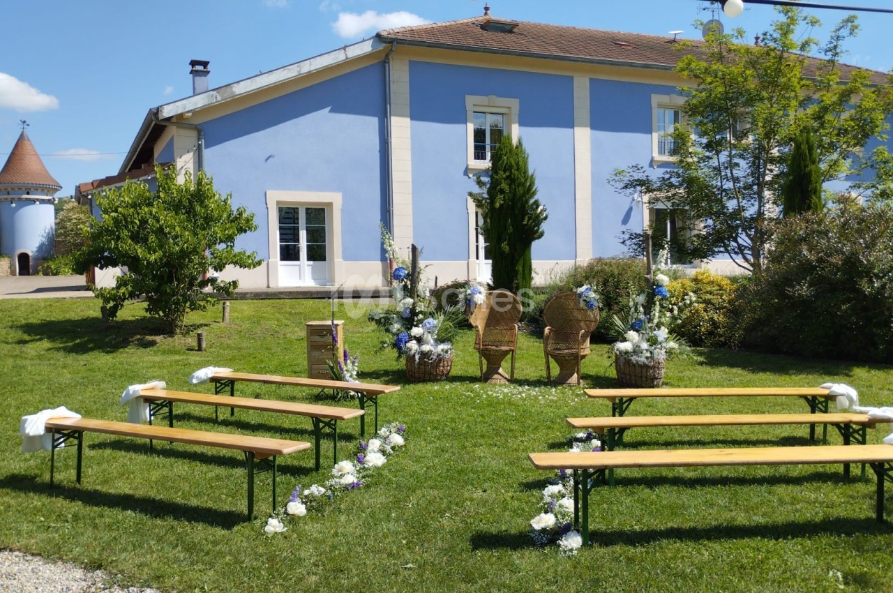 Disposition d'une cérémonie en plein air avec bancs en bois, décor floral et une maison bleue en arrière-plan.