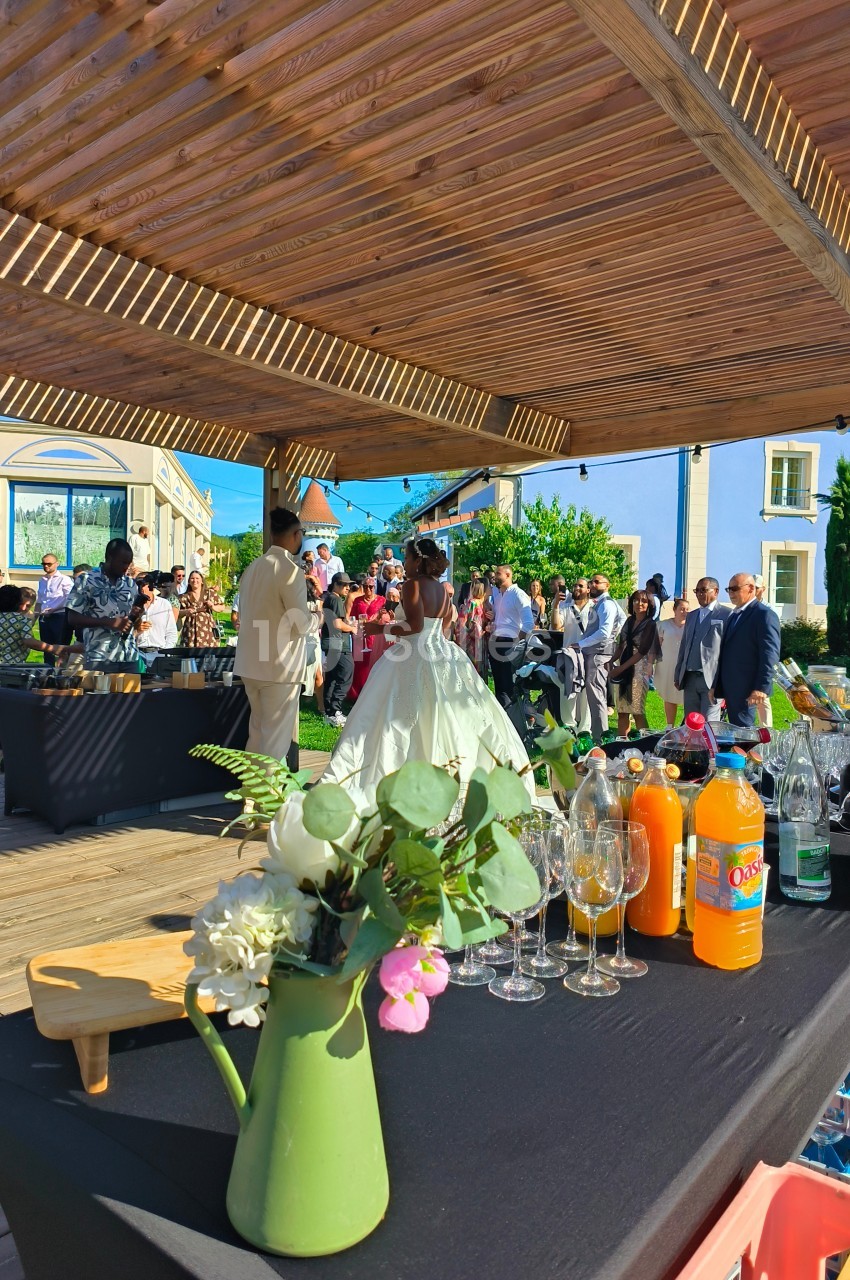 Invités rassemblés sous une pergola en bois lors d'une réception, avec une table de boissons au premier plan.
