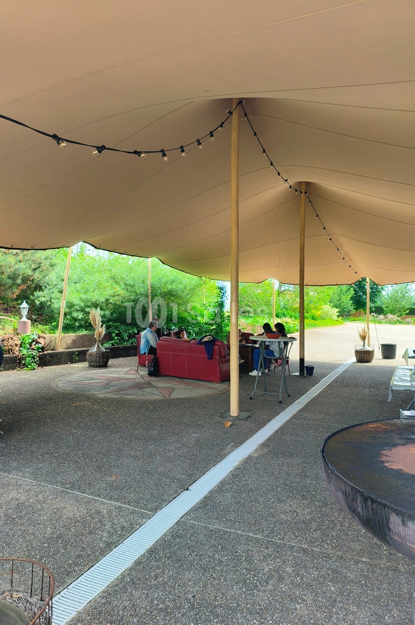 Espace extérieur couvert par une tente beige, avec des tables et des personnes assises dans un cadre verdoyant.