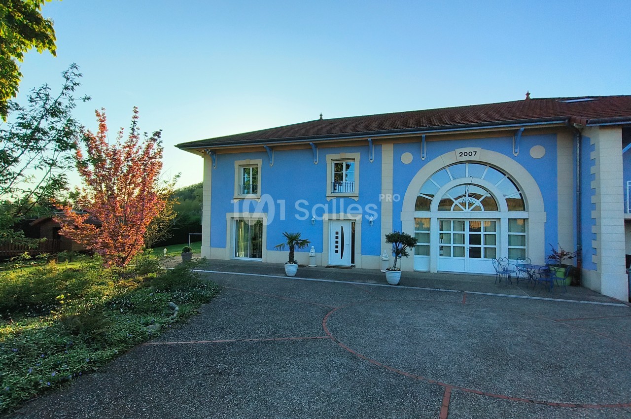 Façade d'une maison bleue avec une grande porte vitrée, entourée de plantes en pot et d'un arbre fleuri.