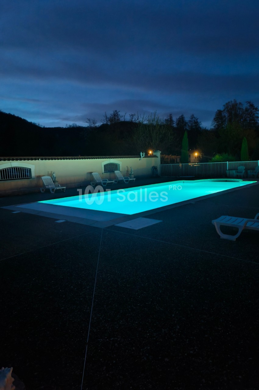 Piscine extérieure éclairée de nuit, entourée de chaises longues et d'arbres en arrière-plan.