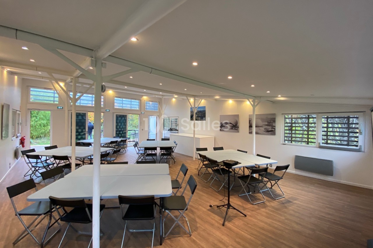 Salle lumineuse avec tables blanches et chaises pliantes, parquet en bois clair et grandes fenêtres laissant entrer la…