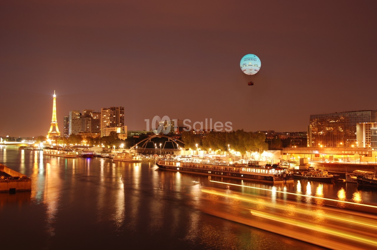 Vue nocturne de Paris avec la Seine, la tour Eiffel illuminée et un ballon captif dans le ciel.
