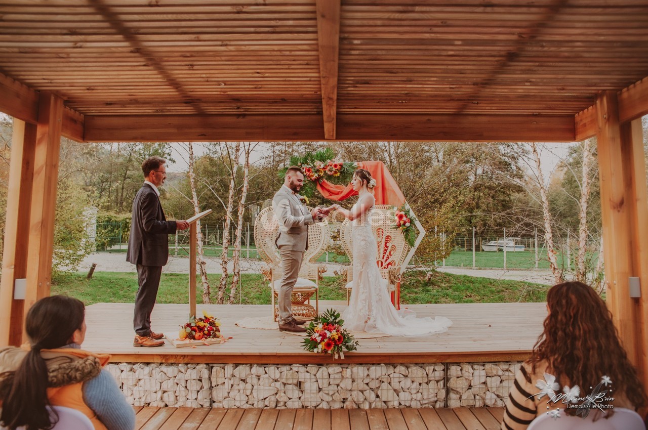 Un couple échange ses vœux de mariage sous une pergola en bois, entouré de fleurs et d'invités.