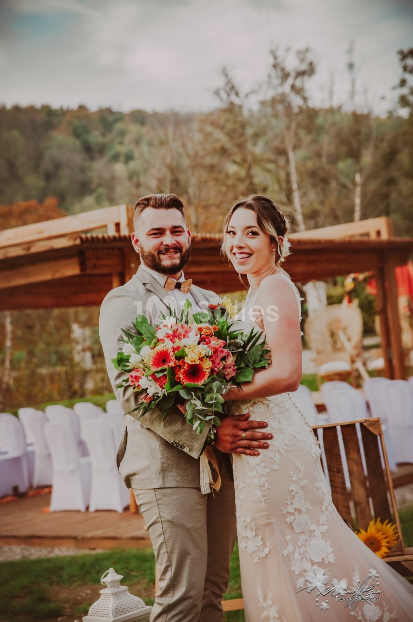 Un couple souriant en tenue de mariage pose devant une décoration extérieure avec des fleurs et des chaises blanches.