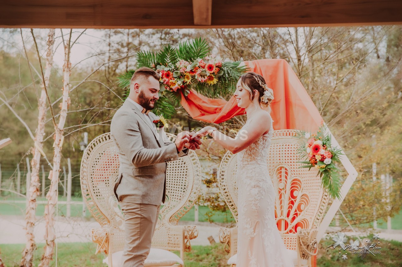 Un couple échange des alliances lors d'une cérémonie en extérieur, devant une décoration florale colorée.
