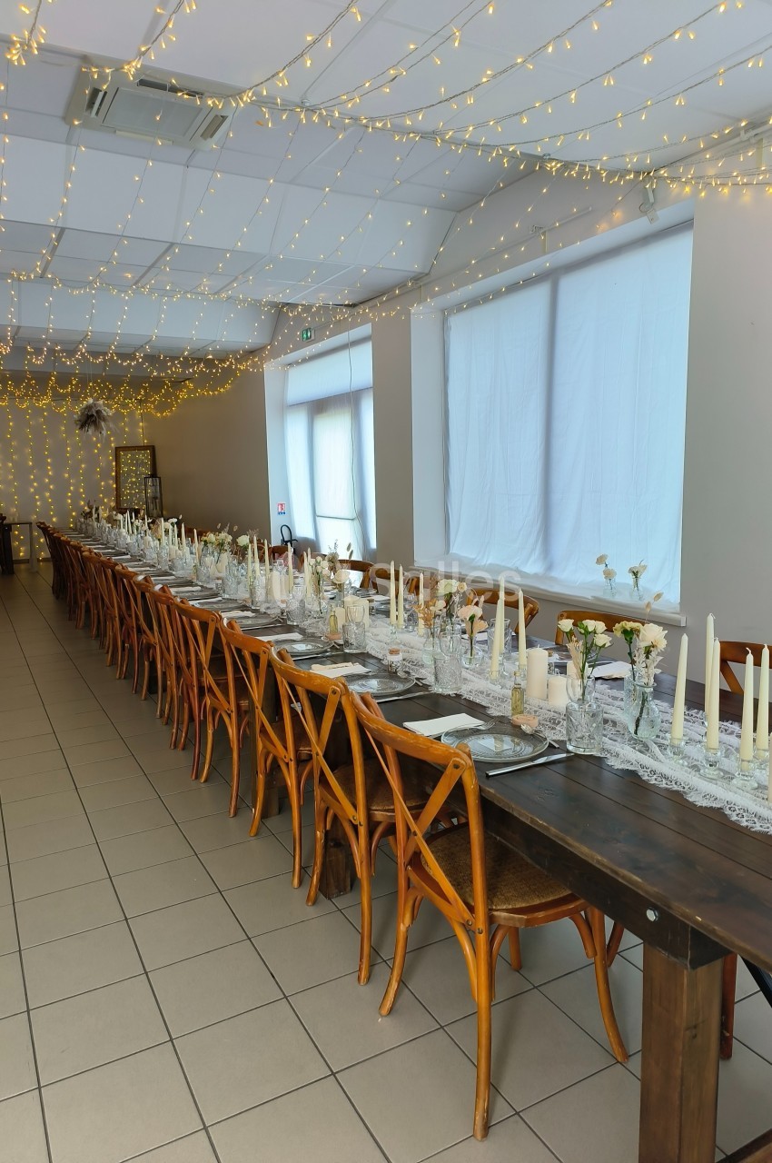 Salle décorée pour un événement avec une longue table en bois, chaises assorties et guirlandes lumineuses suspendues.