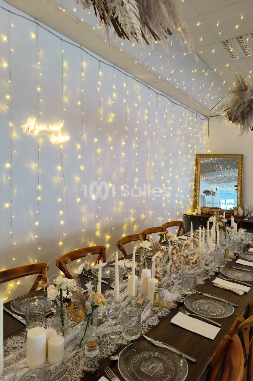 Table décorée pour un événement avec bougies, fleurs, vaisselle élégante et guirlandes lumineuses sur un mur blanc.