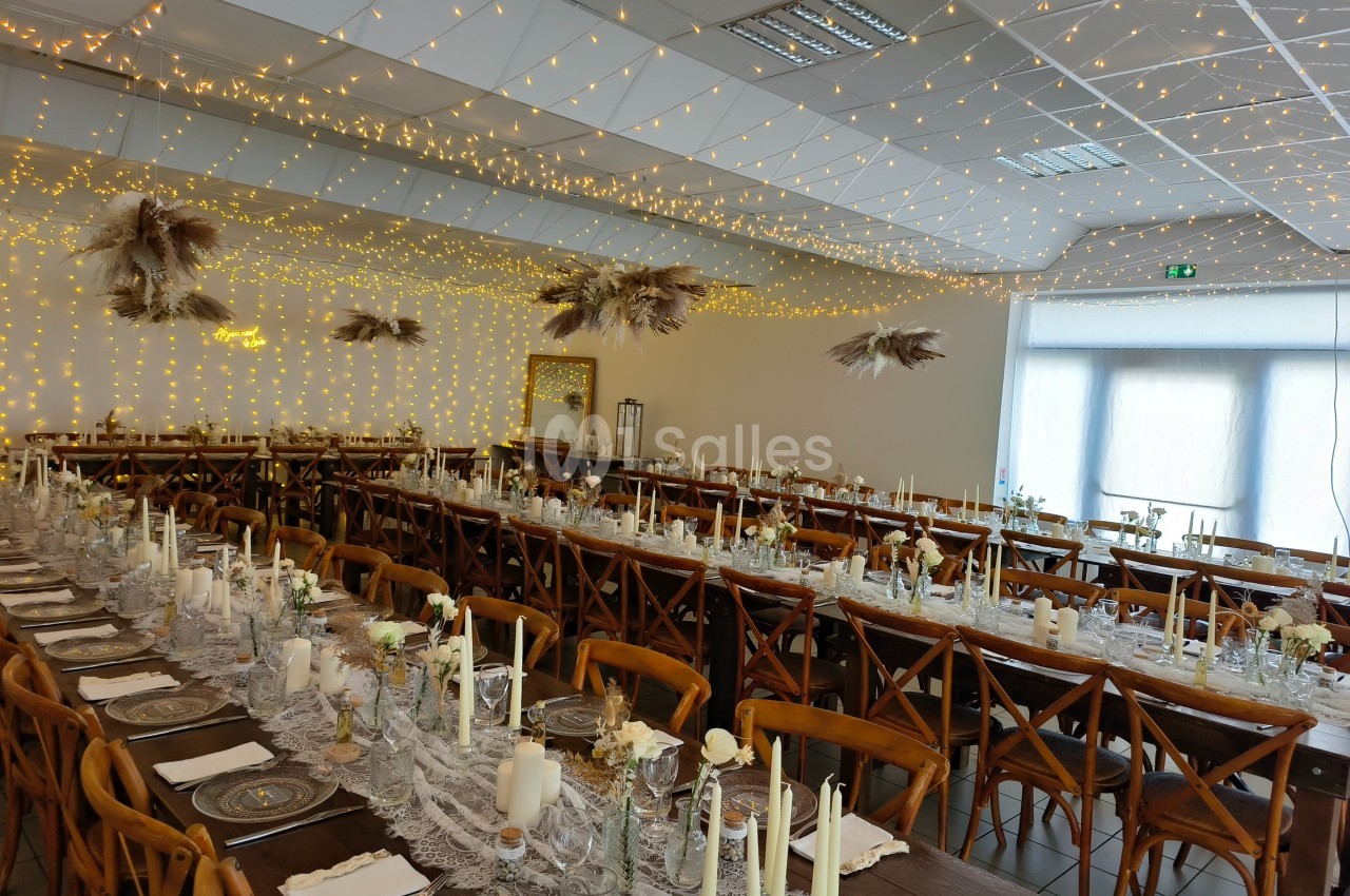 Salle de réception décorée avec des guirlandes lumineuses, tables alignées avec nappes en dentelle et bougies.