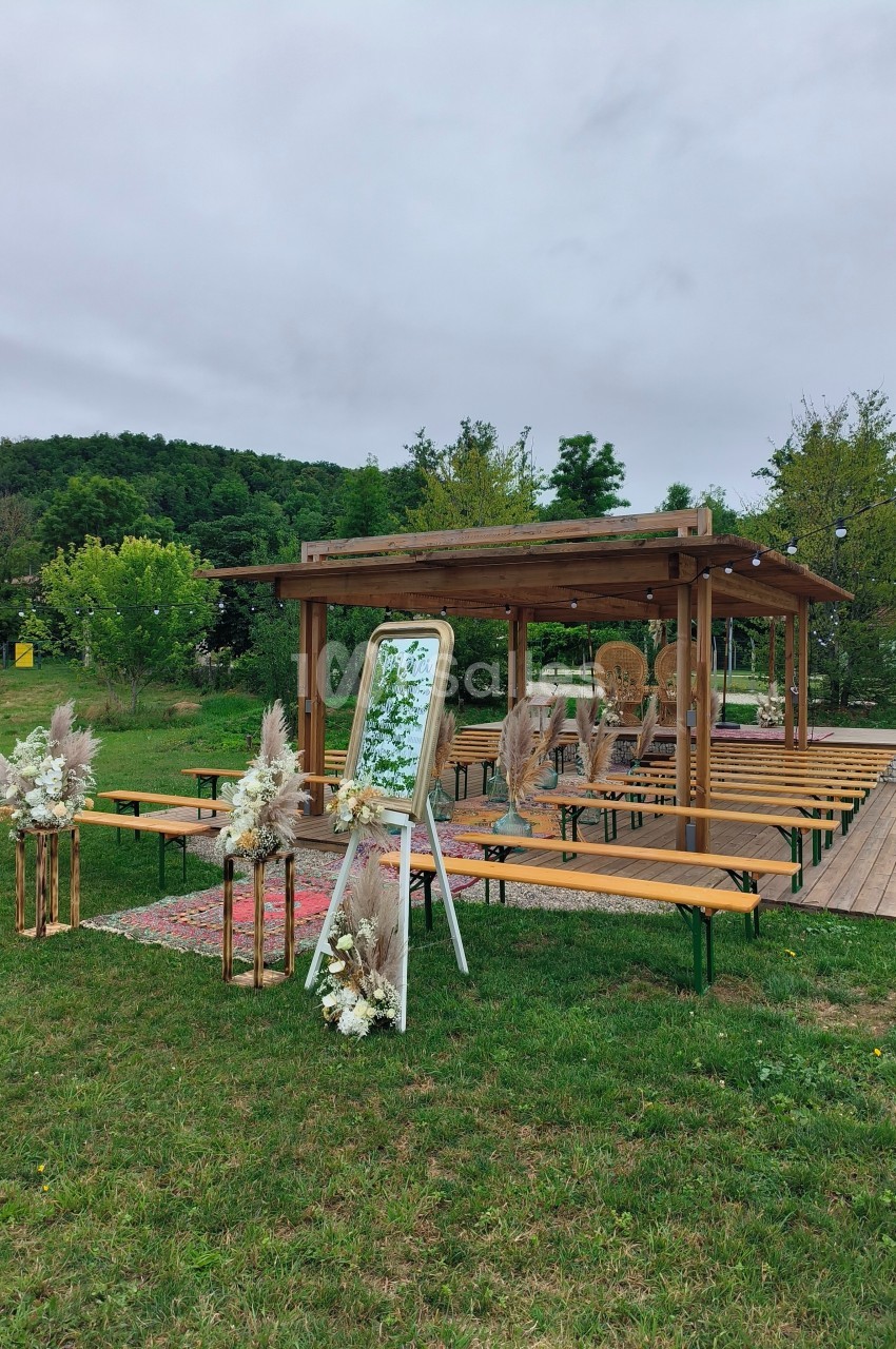Espace extérieur aménagé pour une cérémonie, avec pergola en bois, bancs, décorations florales et miroir.