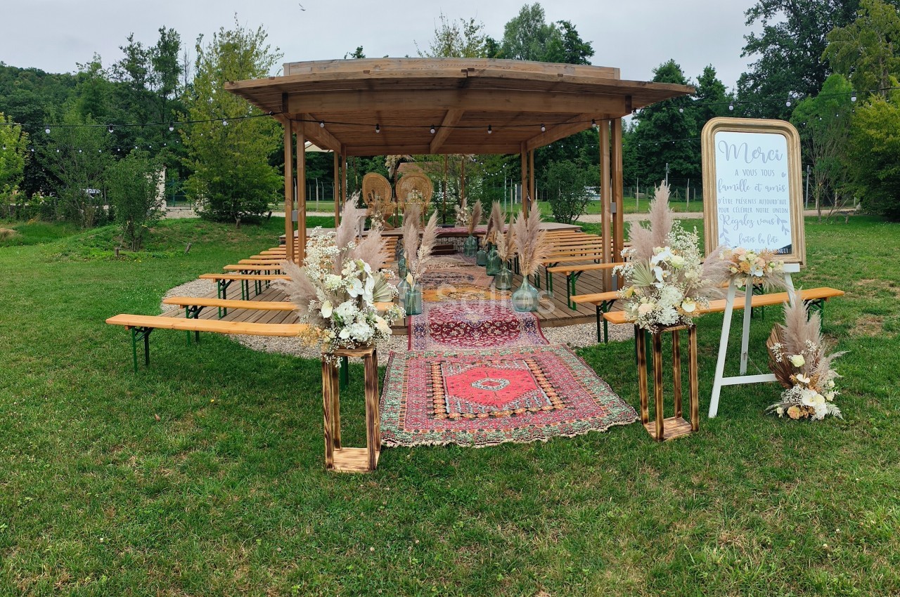 Espace extérieur aménagé avec une pergola en bois, tapis colorés, bancs et décorations florales pour un événement.