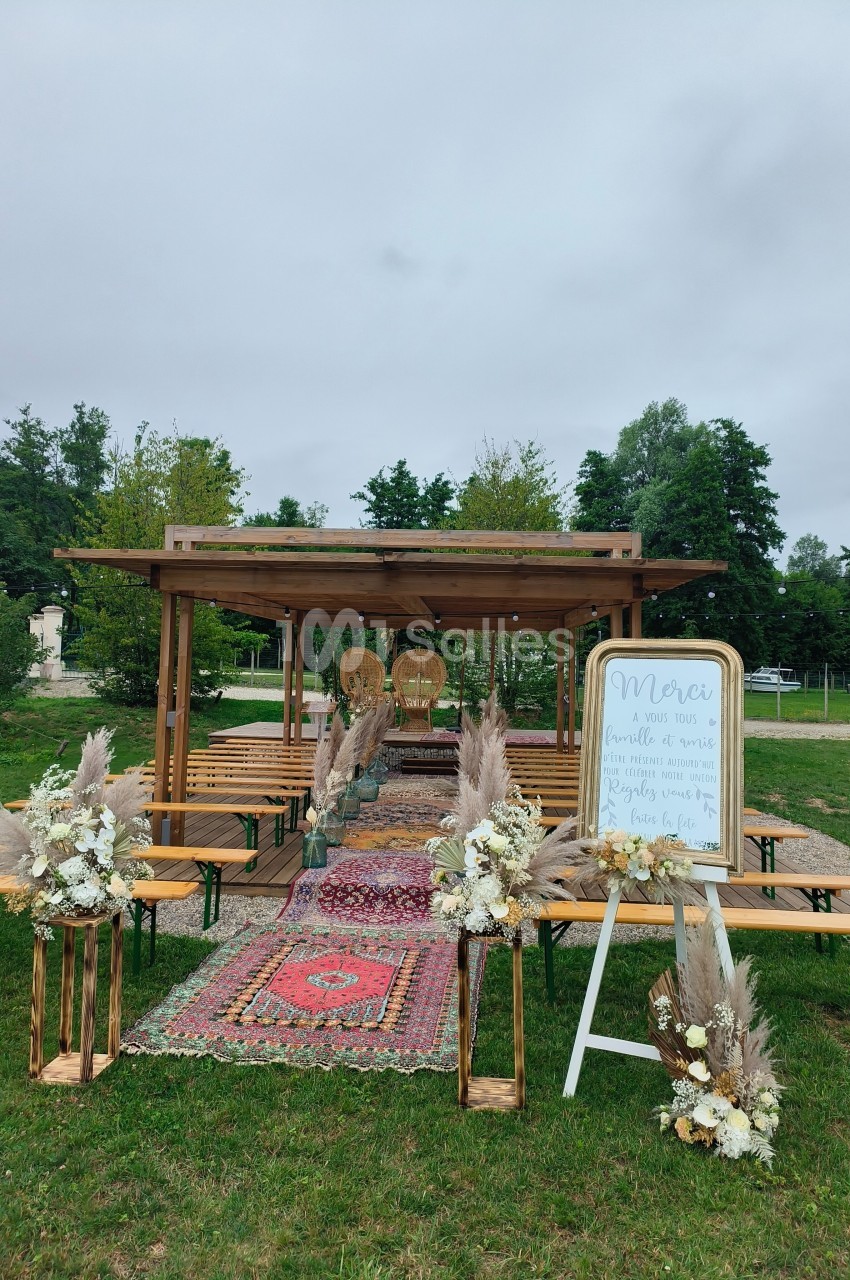 Cérémonie en extérieur avec arche en bois, tapis colorés, bancs en bois et décorations florales.