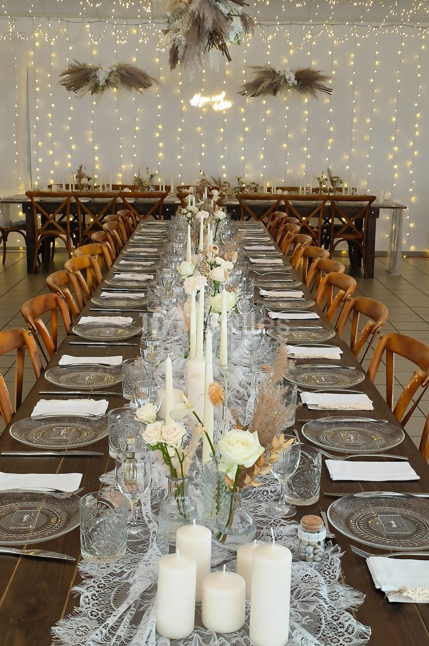 Grande table de réception décorée avec des bougies, des fleurs blanches et des guirlandes lumineuses en arrière-plan.