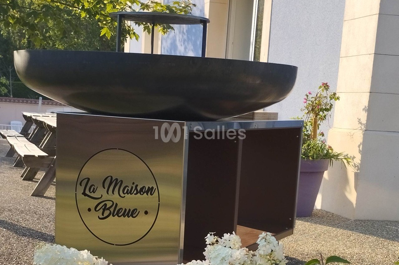 Brasero noir sur socle métallique marqué ’La Maison Bleue’, installé sur une terrasse ensoleillée avec fleurs et tables.