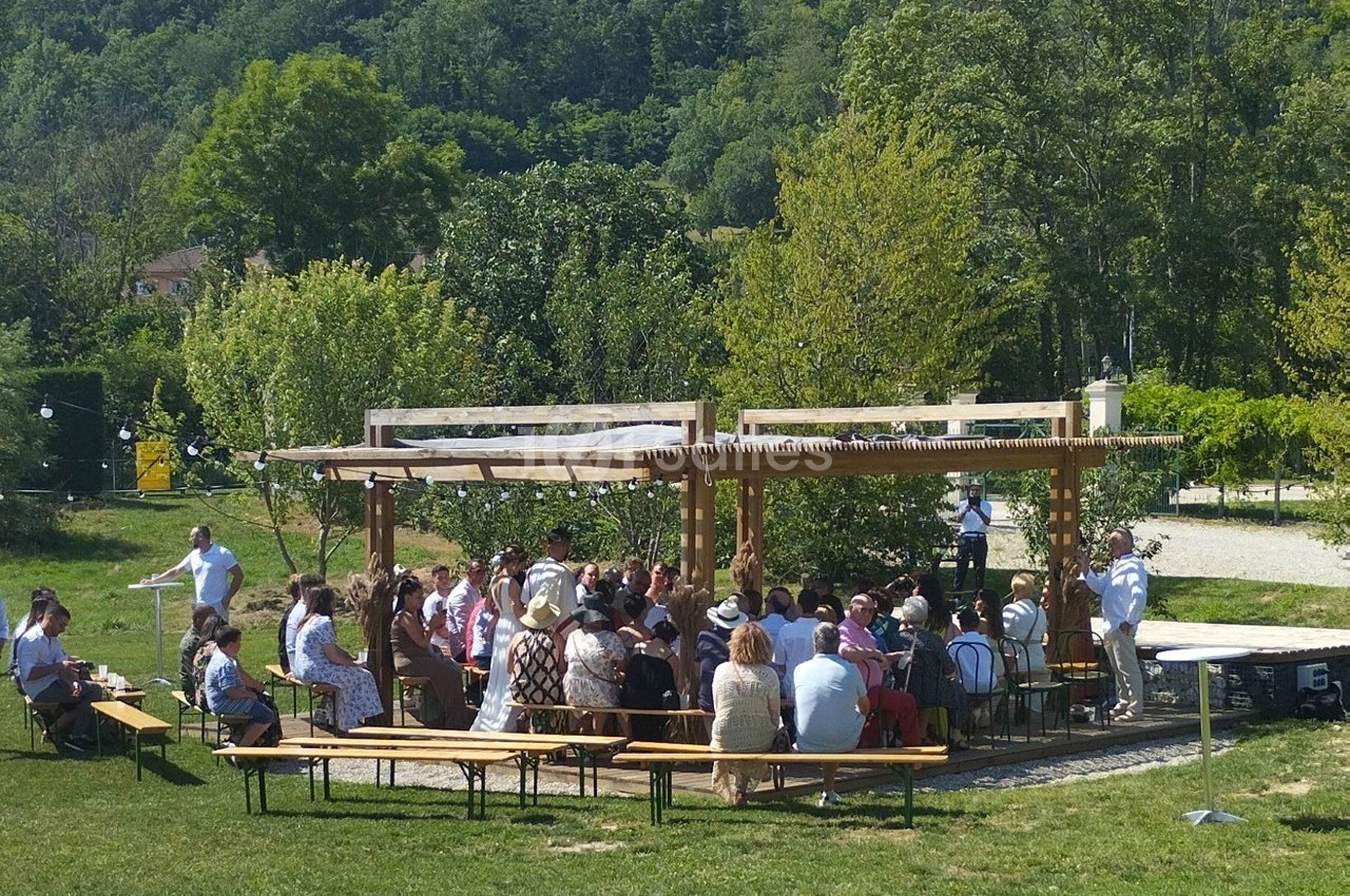 Groupe de personnes assises sous une pergola en bois dans un espace extérieur verdoyant par une journée ensoleillée.