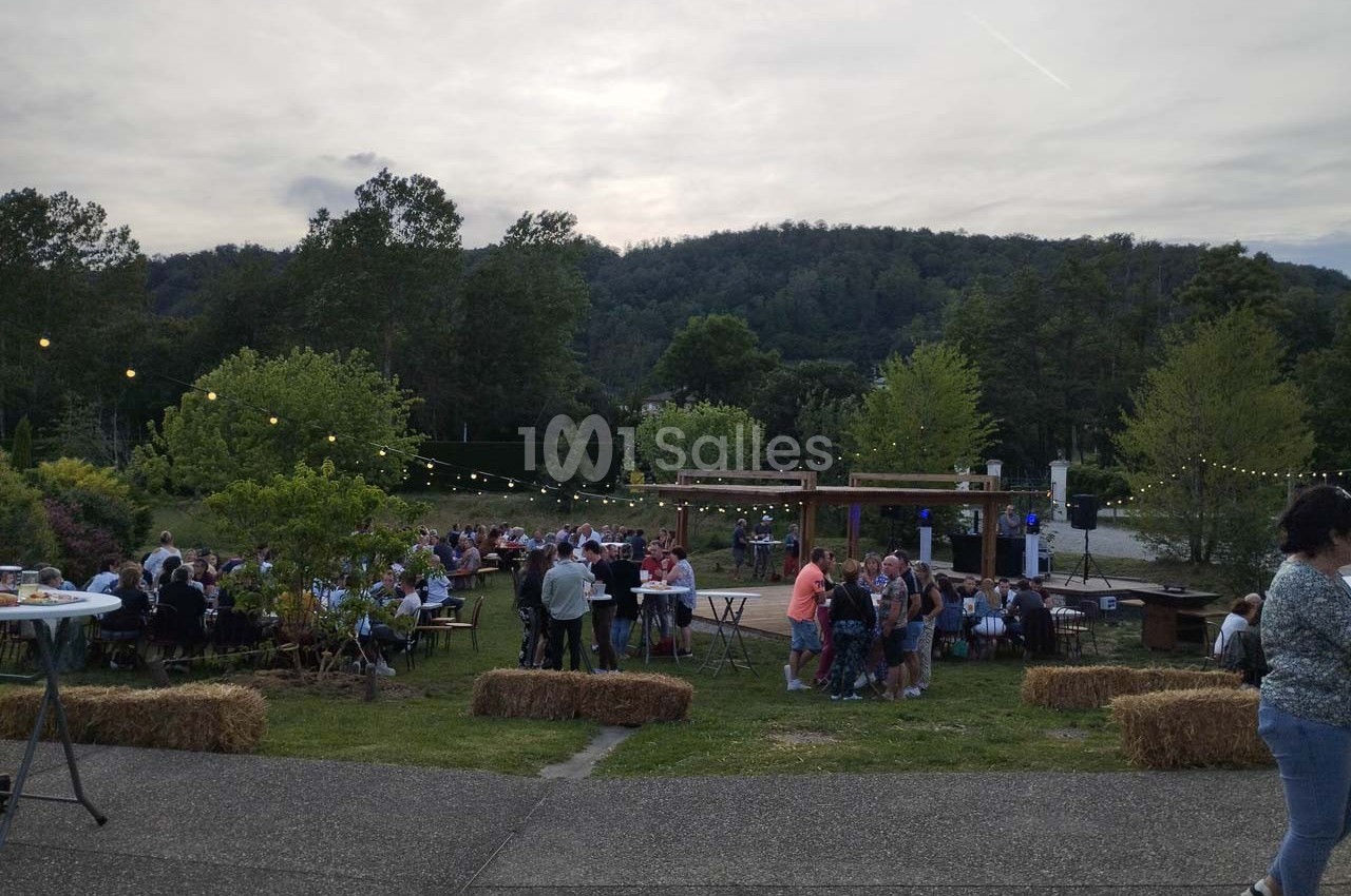 Personnes réunies en plein air autour de tables, avec guirlandes lumineuses et collines boisées en arrière-plan.