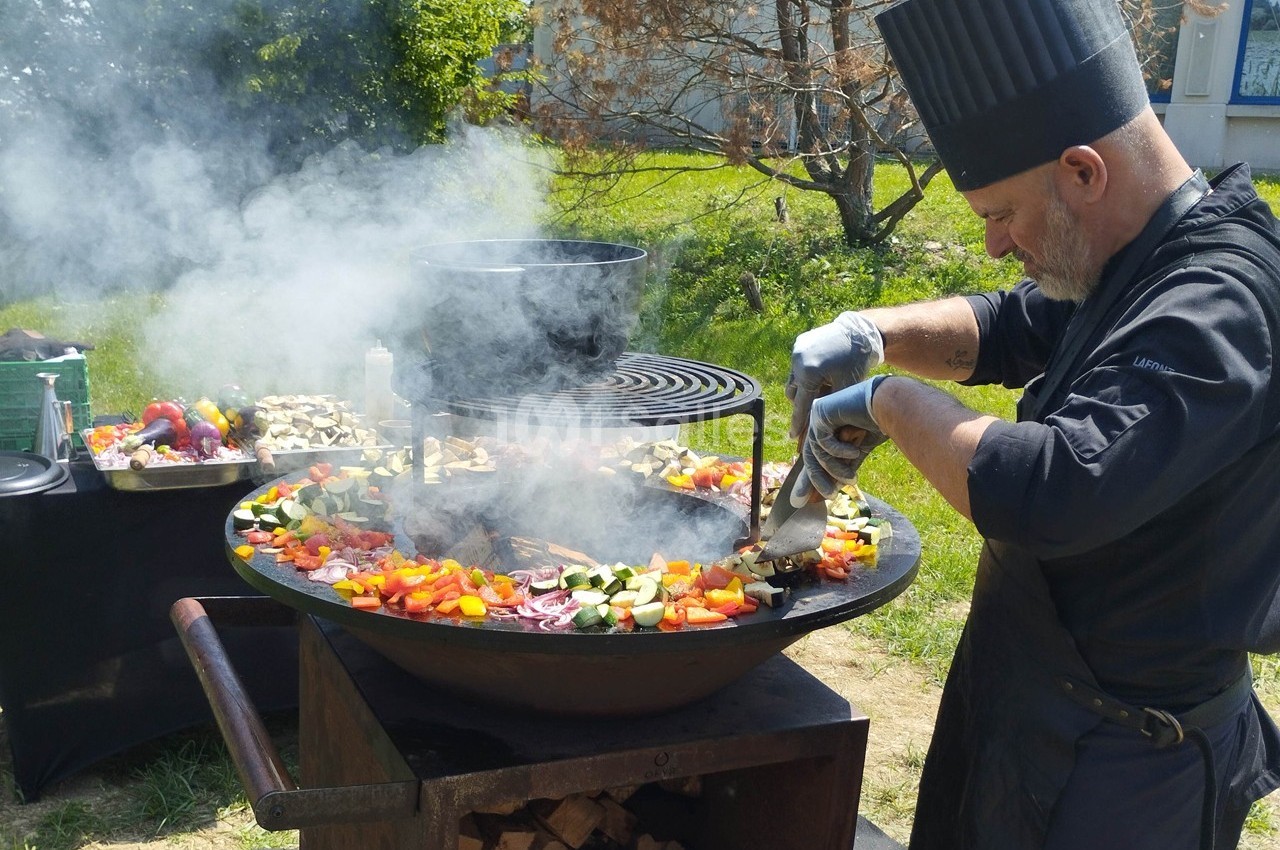 Un chef cuisine des légumes colorés sur une grande plancha en plein air, entouré de fumée et d'ustensiles.