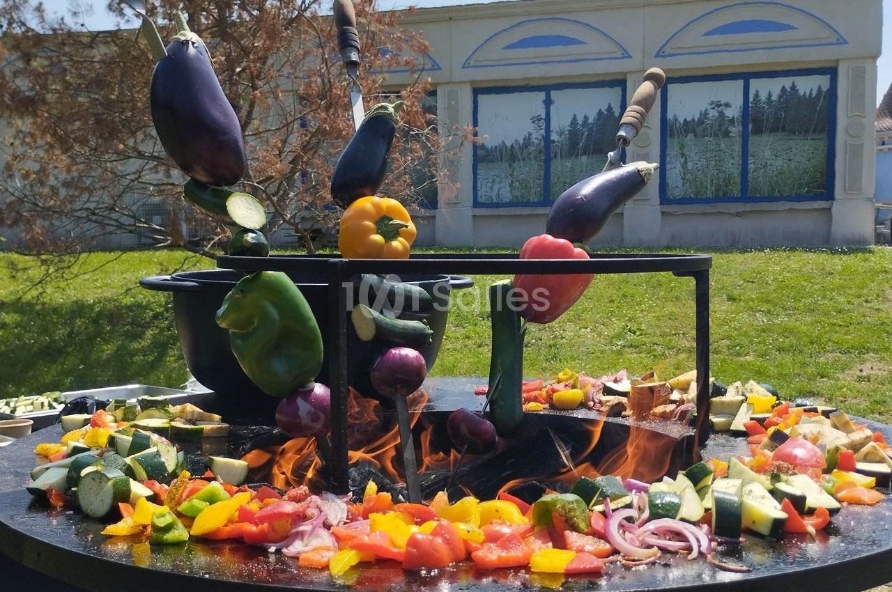 Légumes variés en train de griller sur un barbecue en plein air, avec un bâtiment et de la verdure en arrière-plan.