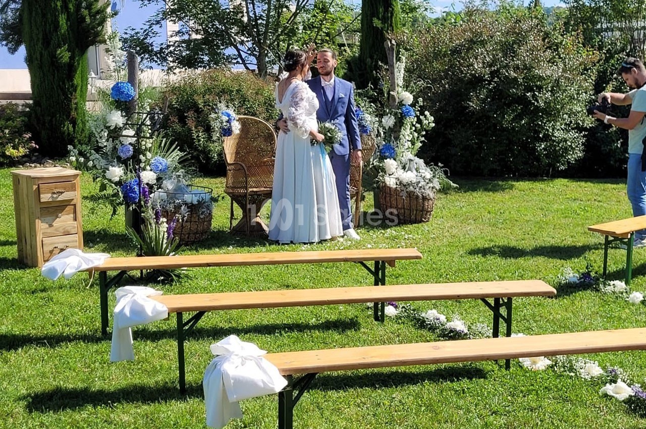 Un couple de mariés debout dans un jardin décoré, entouré de fleurs blanches et bleues, avec des bancs en bois au premier…